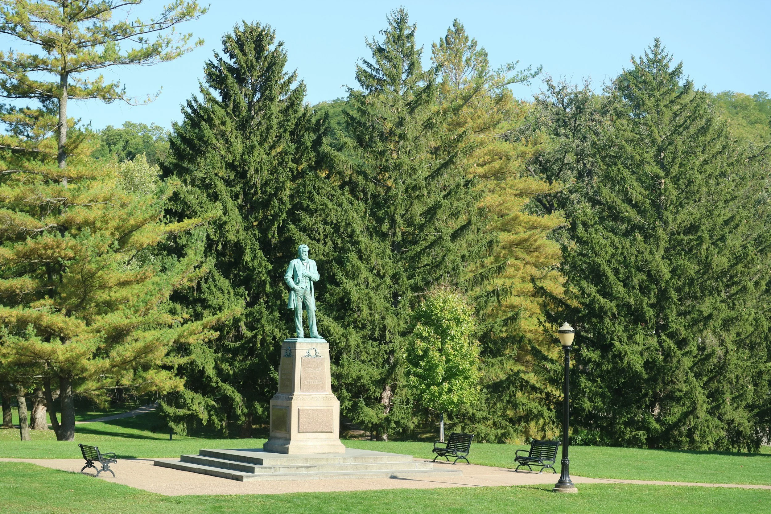 A park scene with a bronze statue of a man standing on a stone pedestal, surrounded by green trees, benches, and a street lamp.