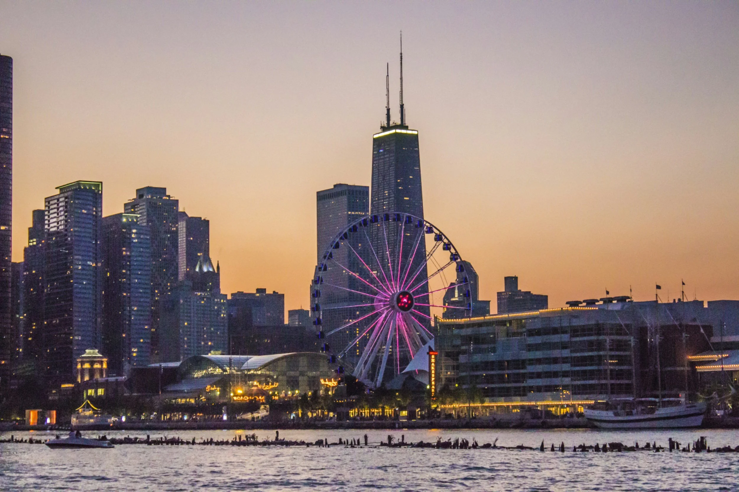 A city skyline at sunset featuring high-rise buildings, a large illuminated Ferris wheel with pink lights, and a body of water in the foreground.
