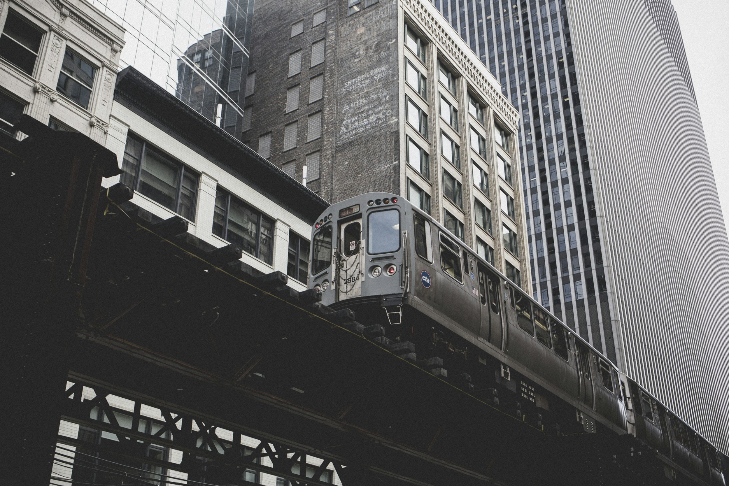 A city scene with an elevated train moving along tracks between tall office buildings, some with glass facades, in an urban environment.