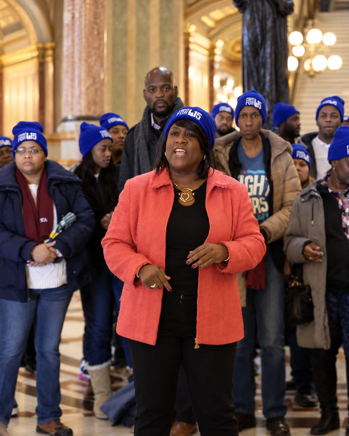 A woman speaking passionately at a rally or event with a large group of people, all wearing blue hats with white writing, in an ornate indoor setting with gold accents.