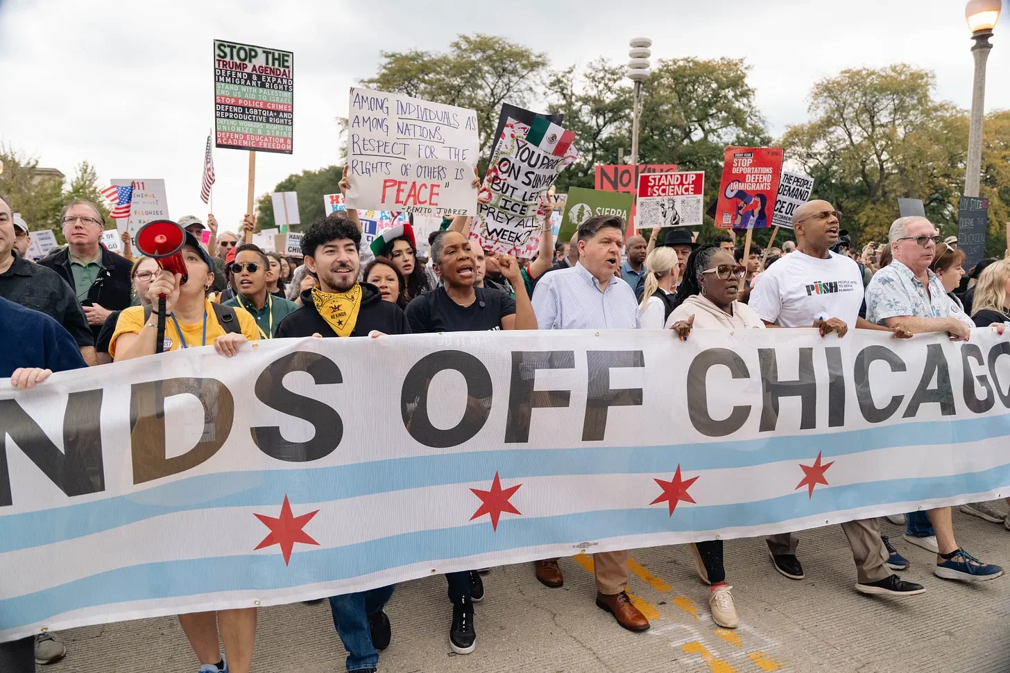 A diverse group of people protesting with signs and a large banner that reads 'KIDS OFF CHICAGO' on a city street with trees and streetlights in the background.