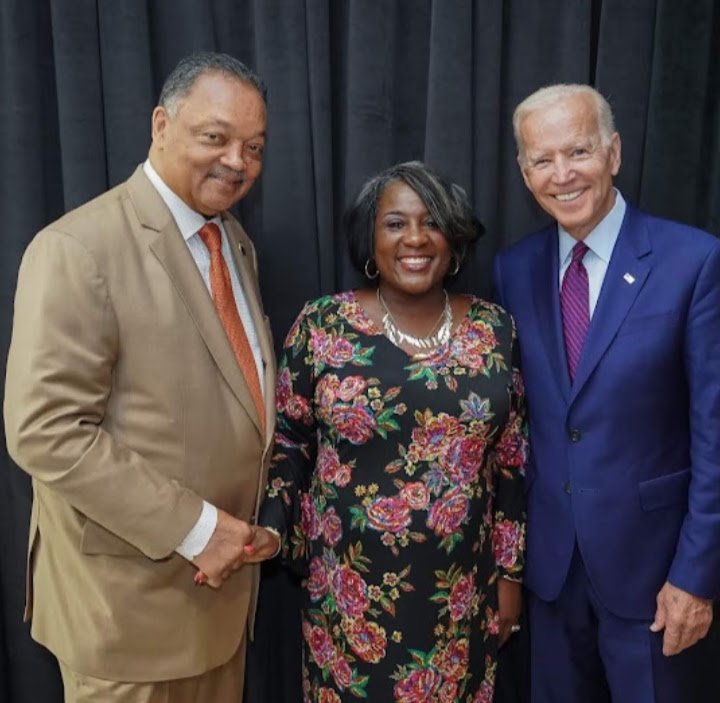 Three people standing together in front of a black curtain, smiling for the photo. The man on the left is wearing a beige suit with a white shirt and a orange tie. The woman in the middle is wearing a dark floral dress with a necklace. The man on the right is dressed in a blue suit with a white shirt and purple tie.