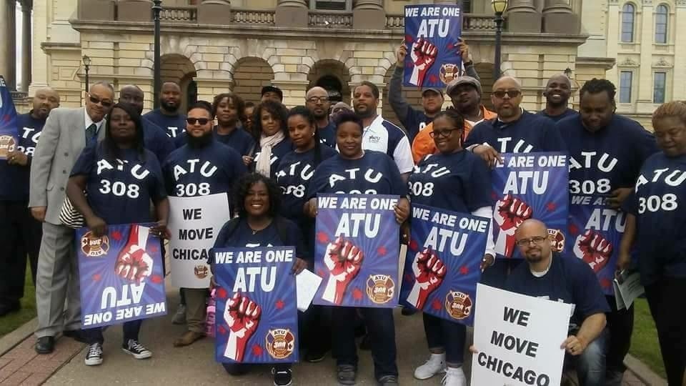 Group of protesters holding signs and banners supporting the American Transit Employees Union (ATU) in front of a historic building. Signs read 'We Are One ATU' with a clenched fist graphic, and others say 'We Move Chicago' and 'We Move'. The participants are diverse, wearing matching blue shirts with 'ATU 308'.