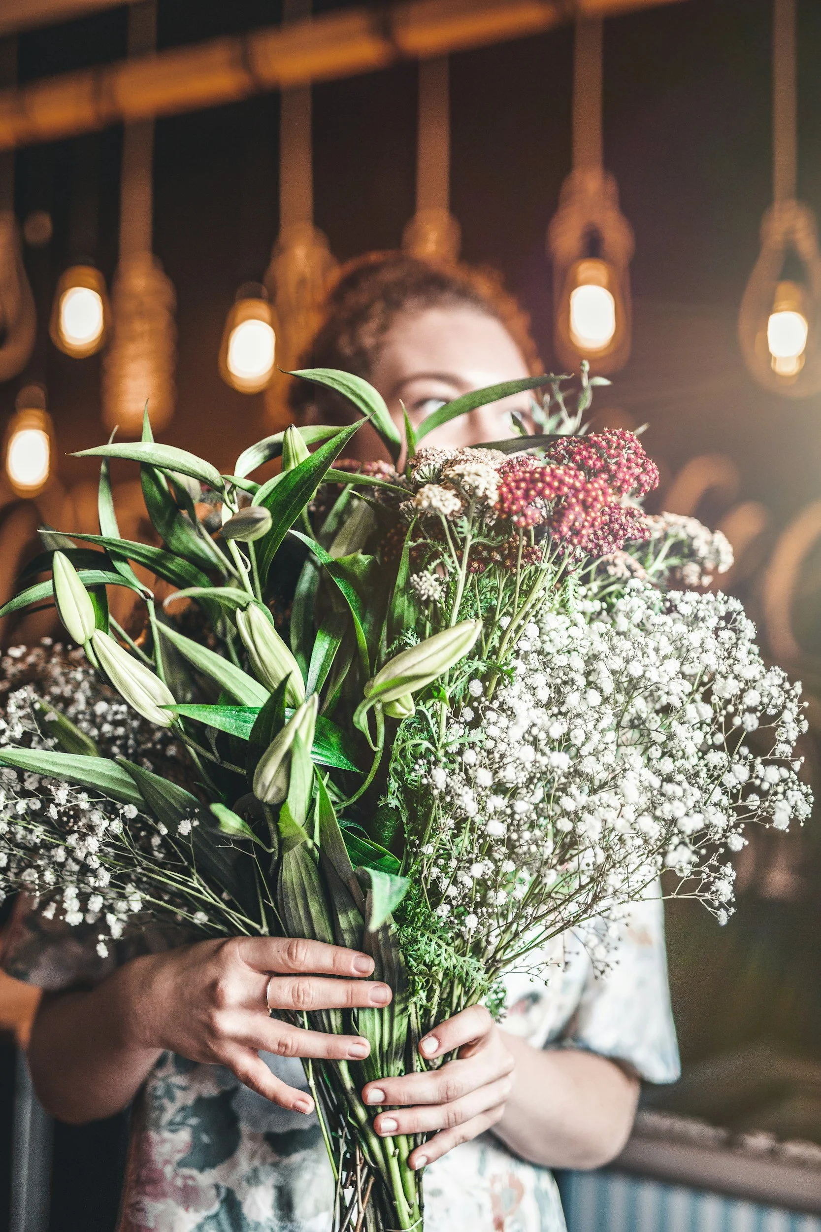 A person holding a large bouquet of mixed flowers, including white baby's breath, lilies, and small red flowers. The person is wearing a floral patterned top, and a warm light hangs in the background.