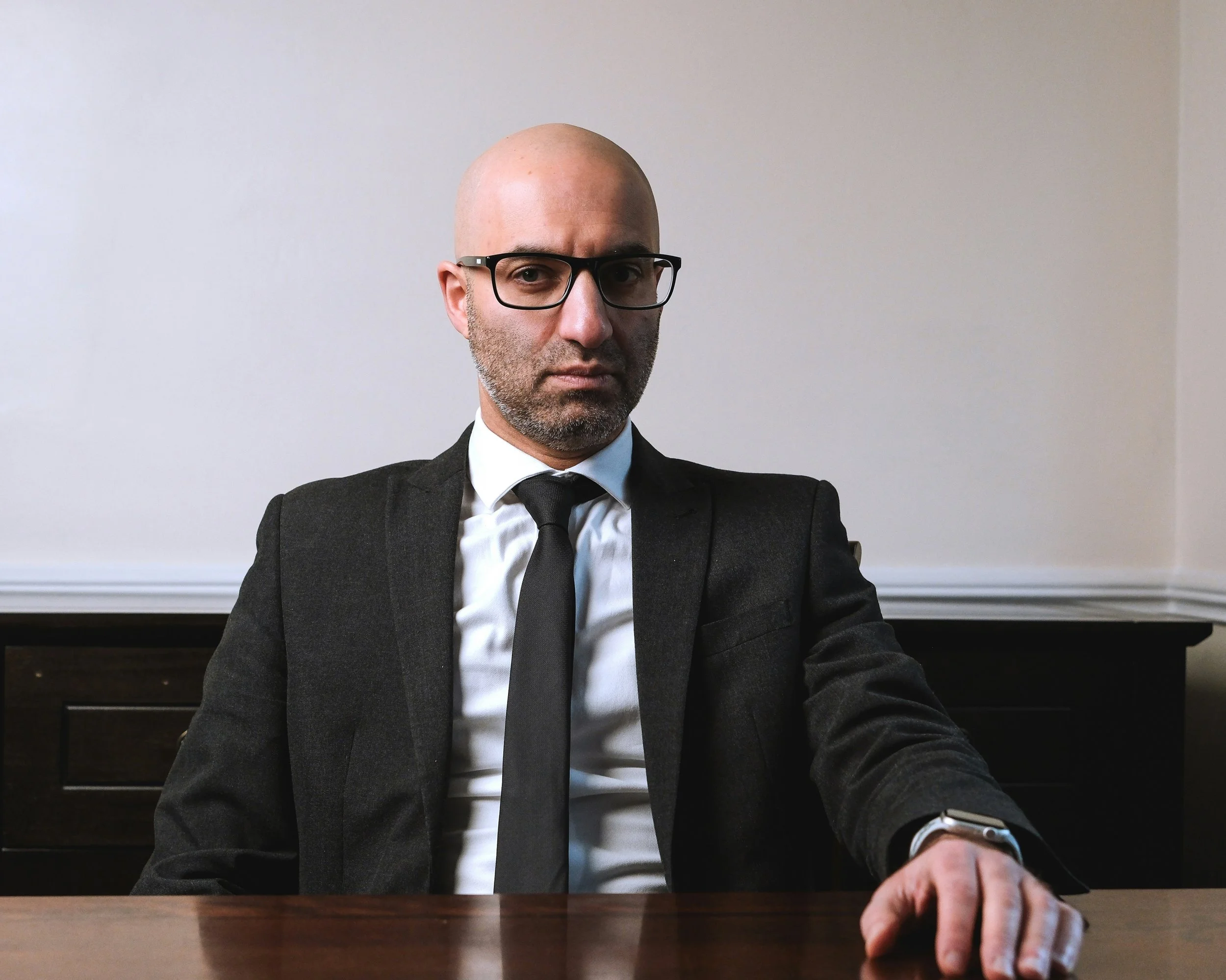 Homme en costume noir, lunettes et cravate noir, assis à une table en bois, regardant directemment l'objectif, dans une pièce avec mur gris clair.