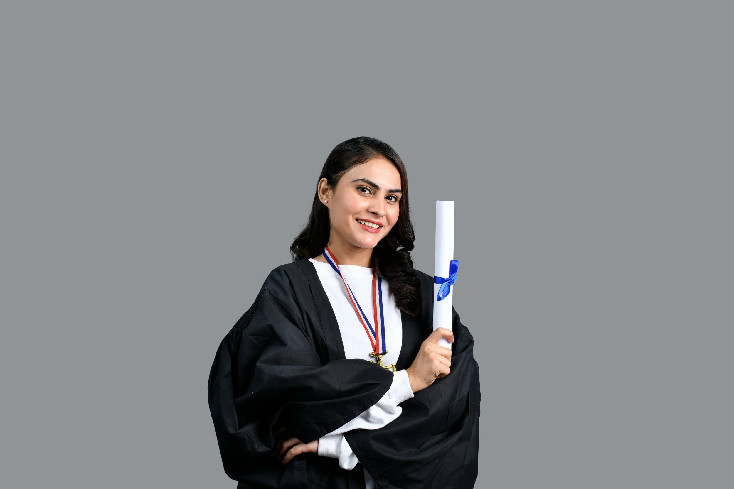 Une femme en robe de diplômée noire, tenant un diplôme roulé avec un ruban bleu, rouge et blanc, souriante, contre un fond gris.