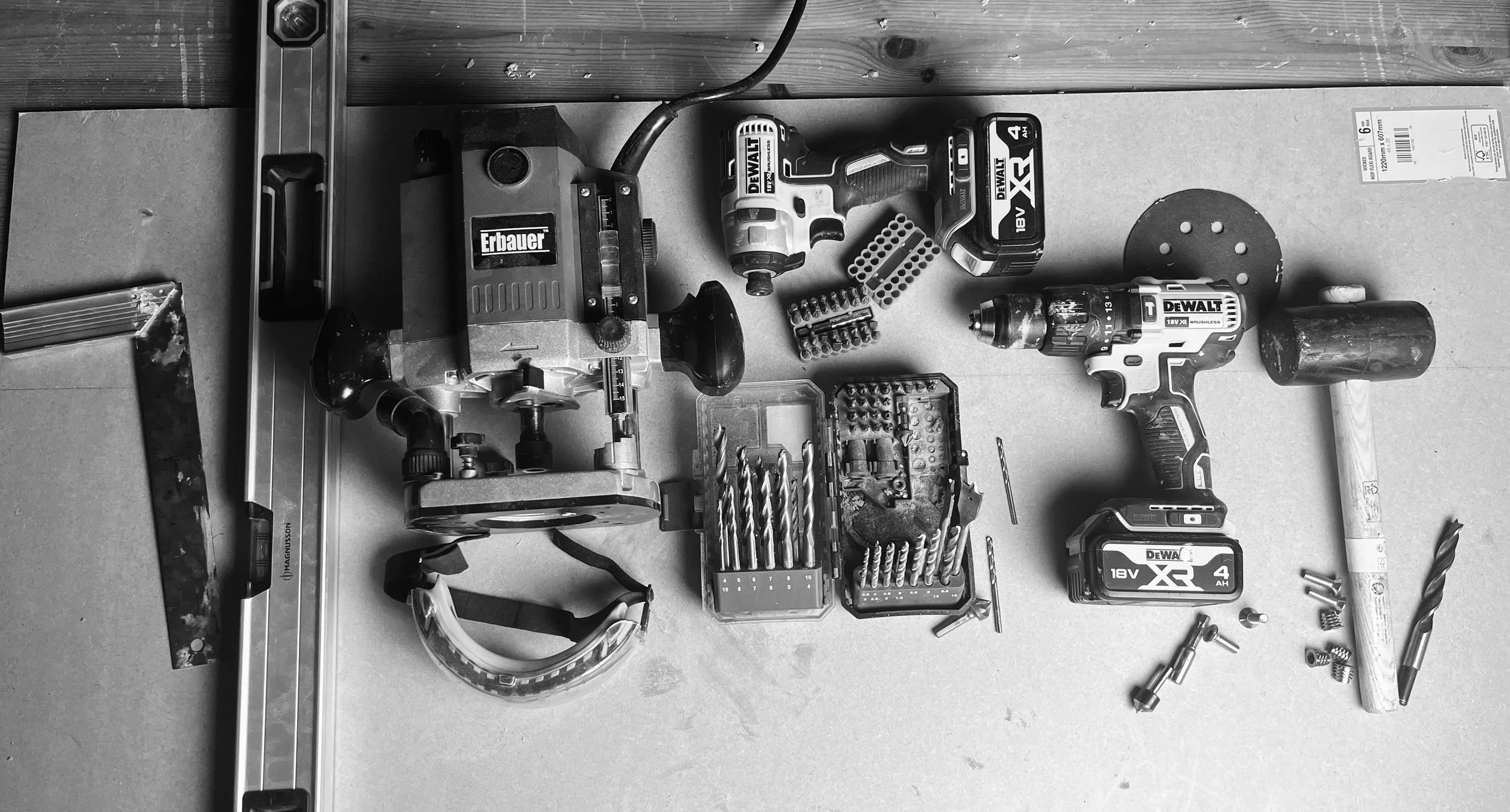 Black and white photo of a workbench with cordless power tools including drills, a saw, and drill bits, along with various screws and hardware.