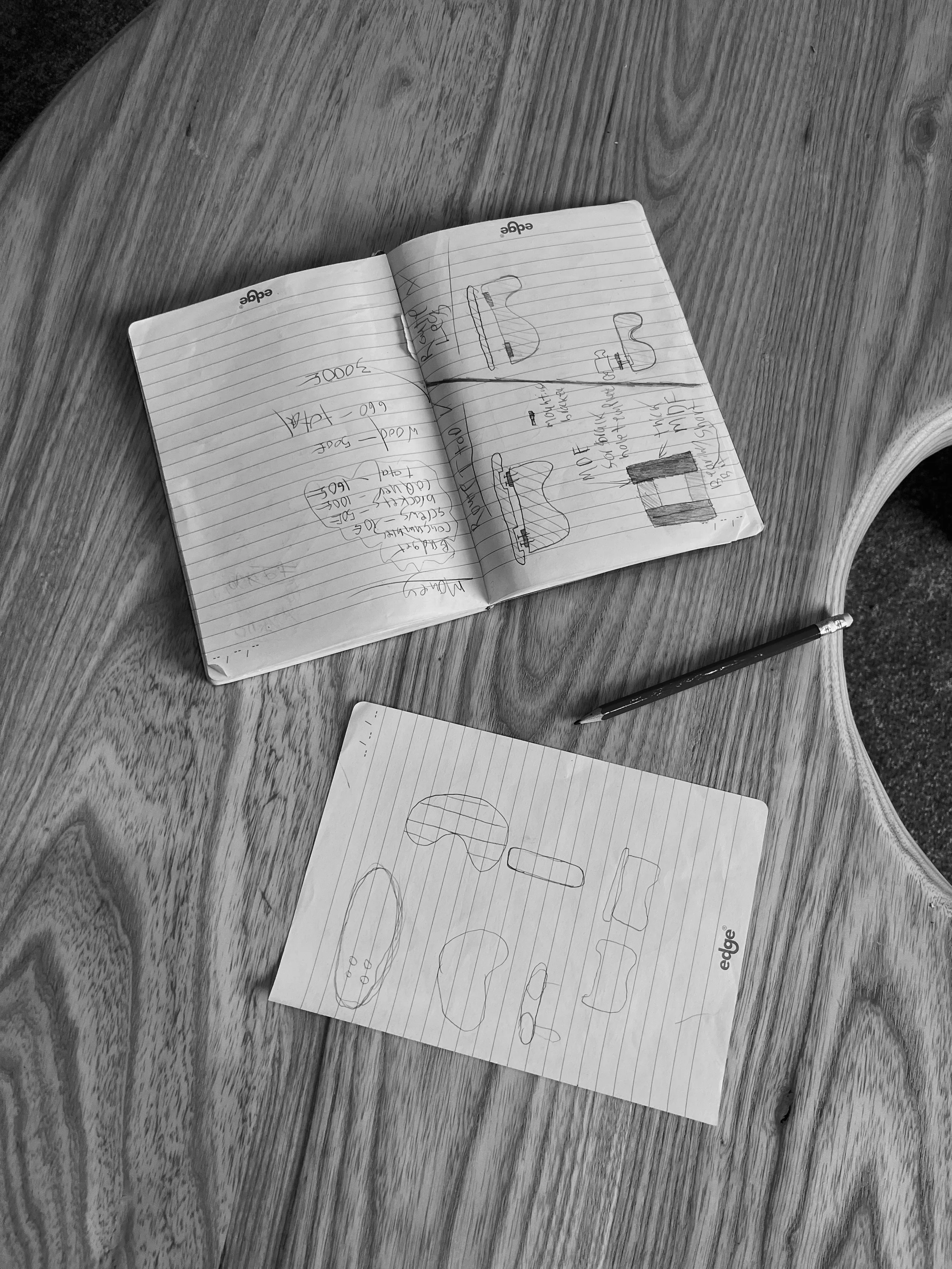 Open notebook on a wooden table with sketches and handwritten notes, with a pen beside it and a sheet of paper underneath.