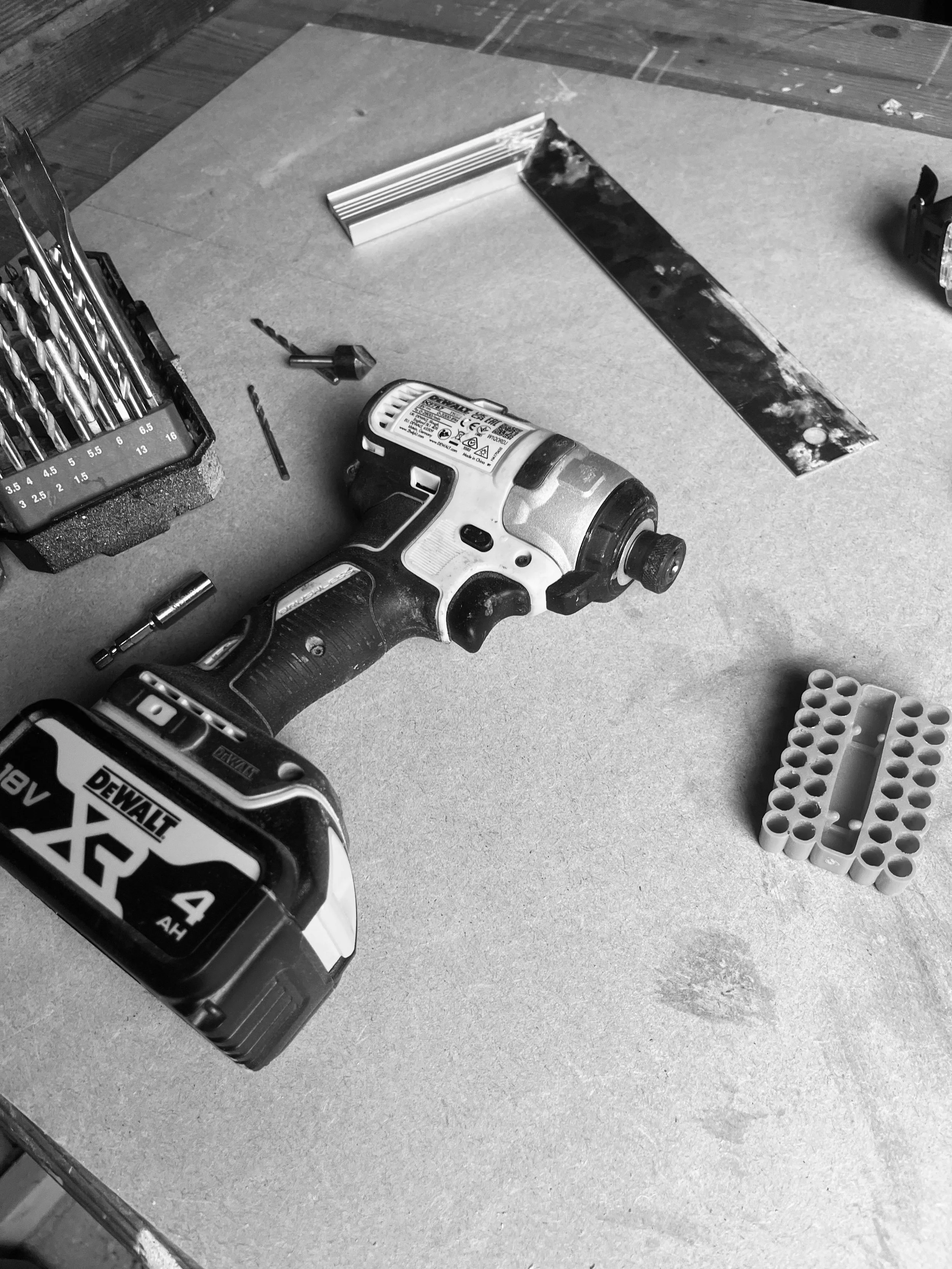 A workbench with a cordless drill, a blade, drill bits, a tape measure, a small container, and a piece of metal or wood with paint or stain on it.