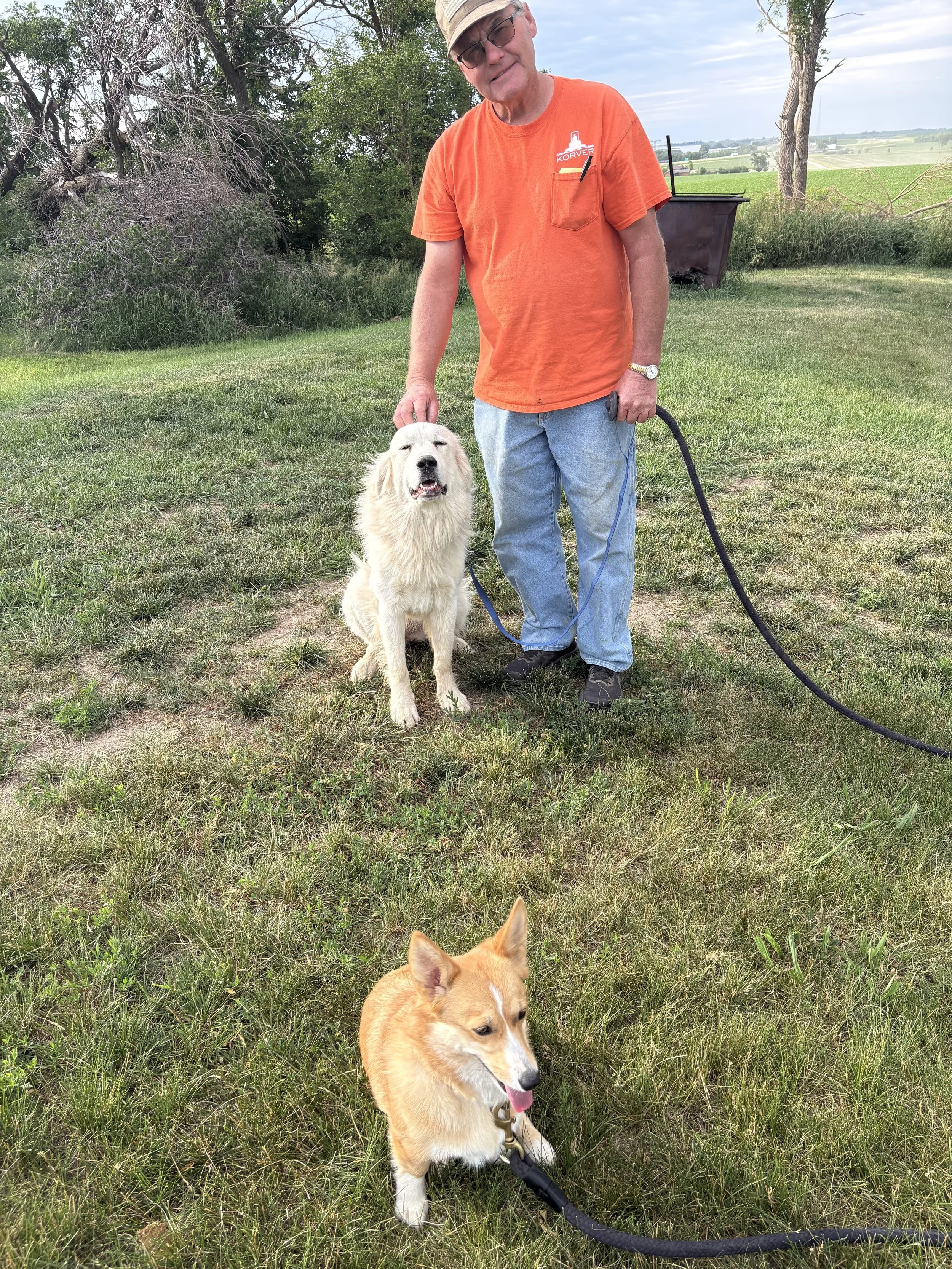 A man wearing an orange shirt, blue jeans, and a beige cap stands outdoors on green grass, holding the leash of a sitting golden retriever. The retriever appears happy with its tongue out and eyes closed. In the background, there are trees, a distant landscape, and a cloudy sky.