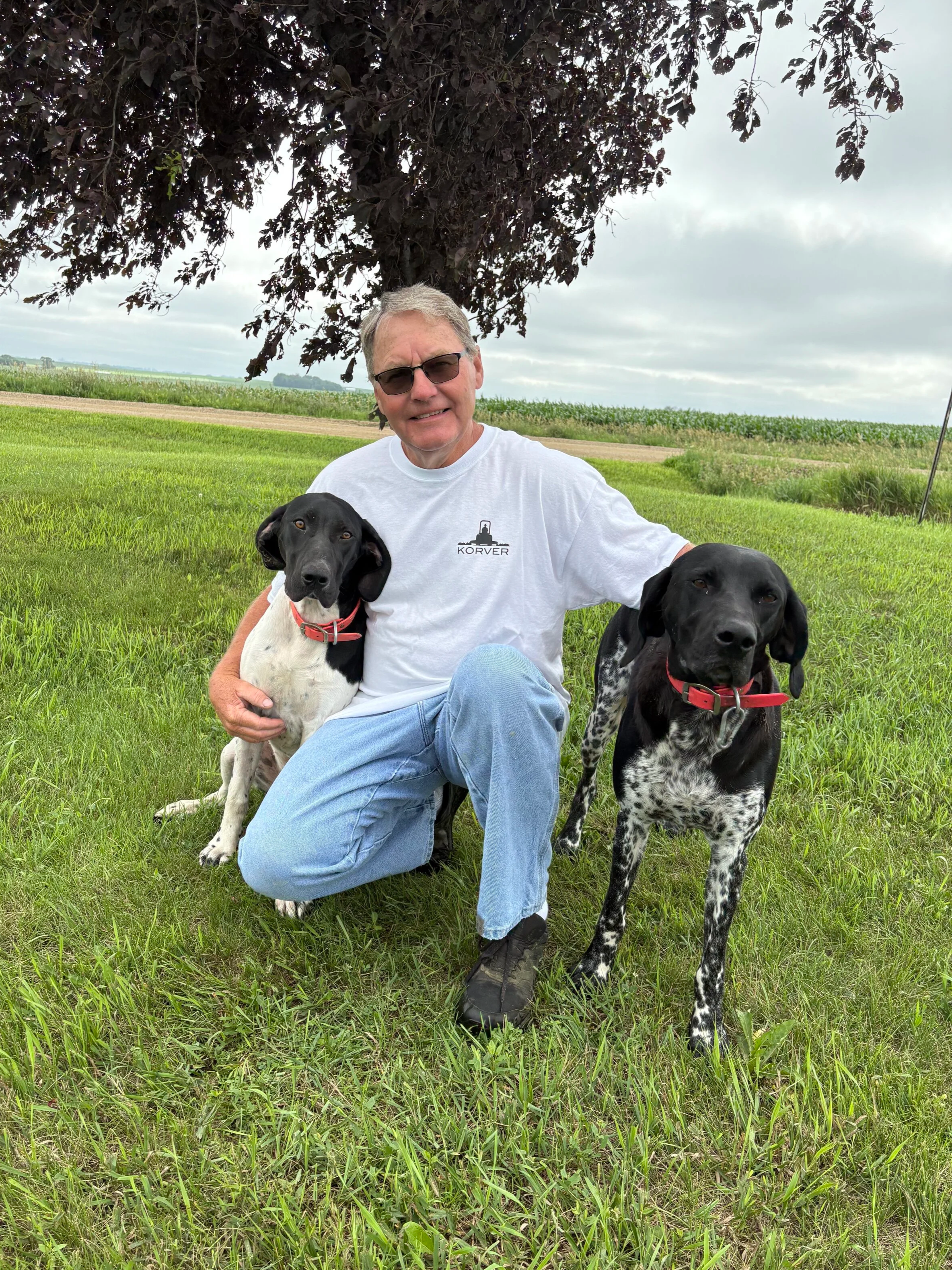 A man sitting on the grass under a large tree with two black and white dogs, one on each side, in a green field with cloudy skies in the background.