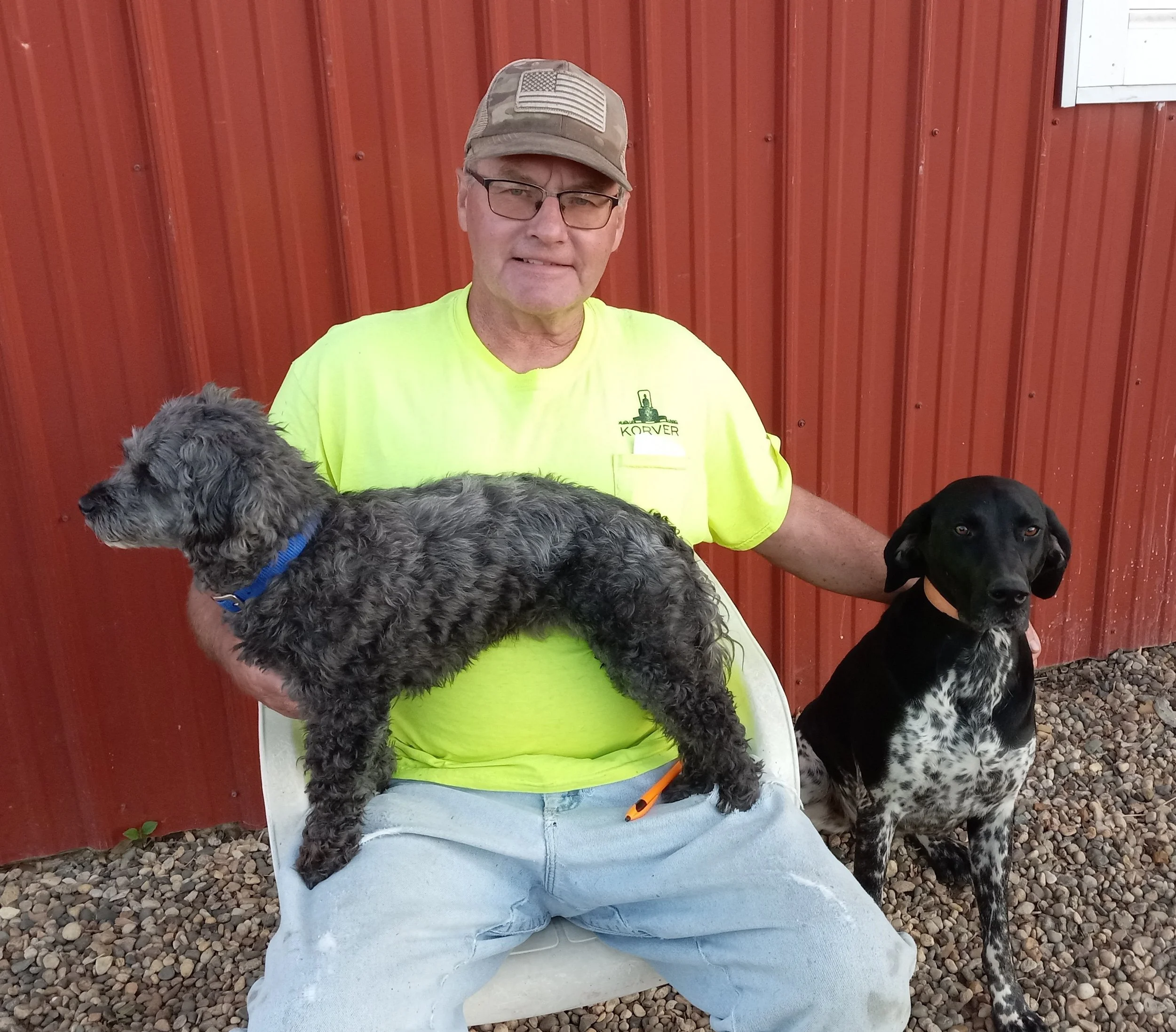 A man wearing glasses, a camouflage American flag cap, and a bright yellow shirt with the word "KORVER" printed on it, sitting on a white chair outside. He is holding a fluffy gray dog on his lap and has a black dog beside him. The background features a red metal wall and gravel ground.