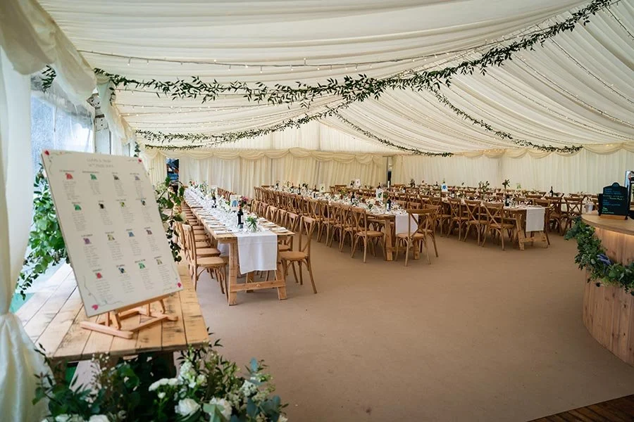 Elegant event tent with long, white-draped tables and wooden chairs, decorated with greenery and candles, set up for a celebration or wedding reception.