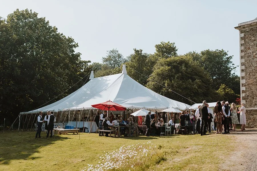 A large white event tent set up outdoors surrounded by tables, chairs, umbrellas, and people attending a gathering on a grassy area with trees in the background.