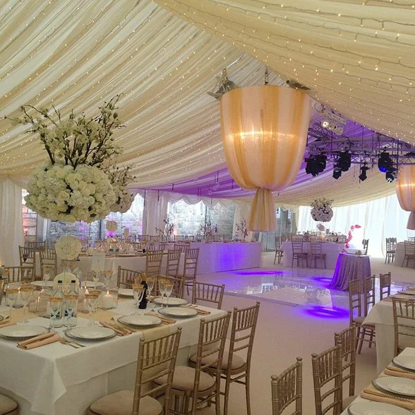 Elegant event tent decorated with draped fabric, hanging floral arrangements, and large illuminated golden centerpieces, with round tables set for a celebration.