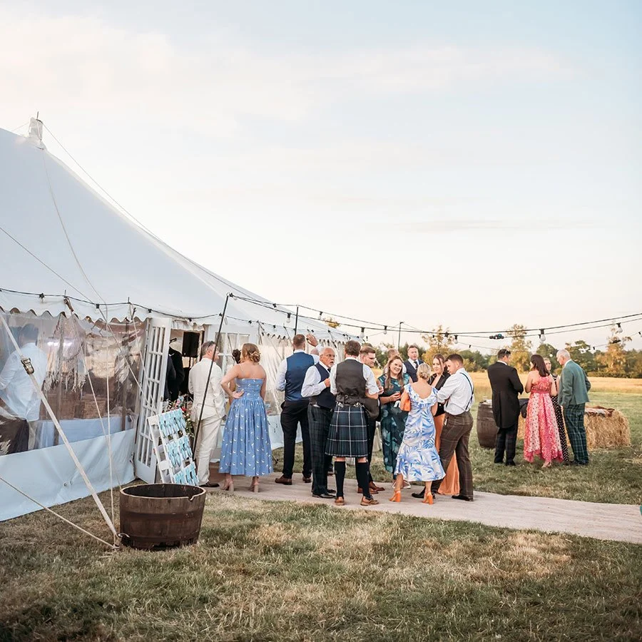 People dressed in formal and semi-formal attire are gathered outdoors near a large white event tent, enjoying a social event or celebration during the daytime.