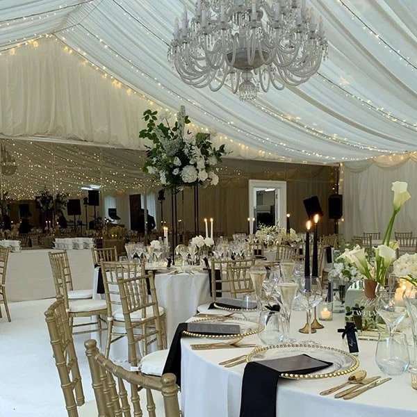 Elegant event setup inside a decorated white tent with chandeliers, string lights, and a large floral centerpiece, featuring round tables with white tablecloths, gold chairs, and black napkins.