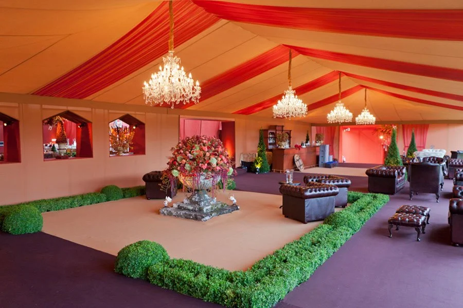 Decorated event tent with chandeliers, floral arrangements, and seating area with armchairs and ottomans.