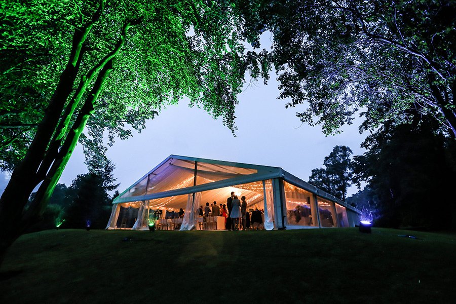 A large white event tent with transparent sides, illuminated from inside, set up on a grassy area surrounded by trees, with people gathered inside during nighttime.
