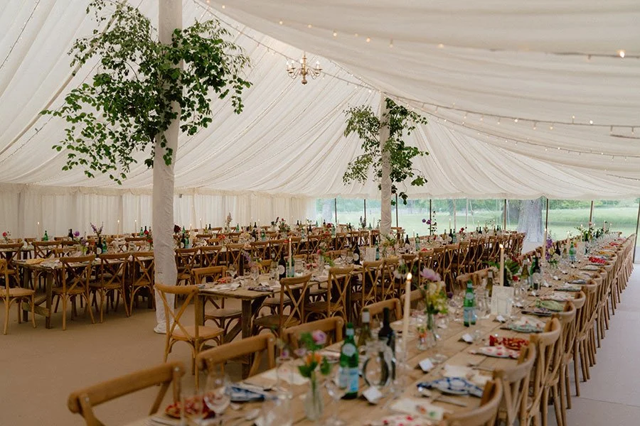 Wedding reception setup inside a decorated tent with long wooden tables, chairs, floral centerpieces, and string lights.