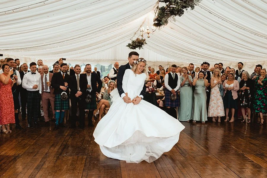 A bride and groom dancing at their wedding reception with guests watching and celebrating in a decorated tent.