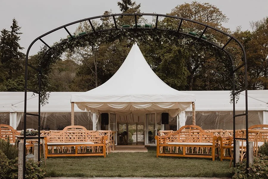 Outdoor event setup with a white tent, wooden benches, and a metal archway, surrounded by trees.