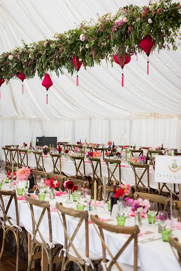 Elegant wedding reception tent with long tables decorated with pink floral centerpieces, green glasses, and place settings. A floral and greenery chandelier with pink lanterns hangs from the ceiling.