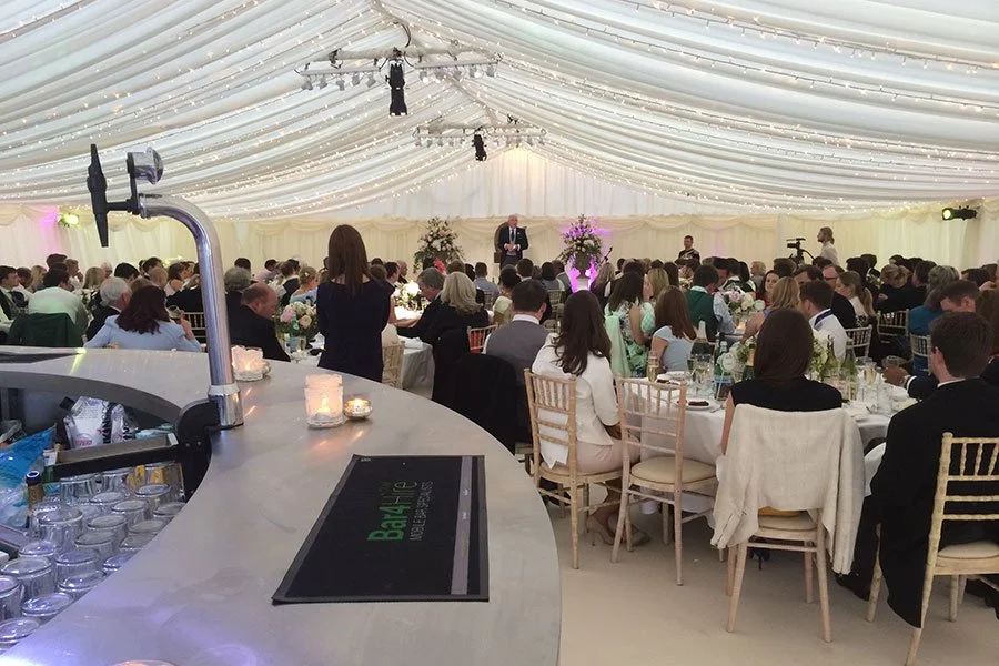 Guests seated at round tables during a formal event in a decorated large tent, with a speaker on stage at the front.