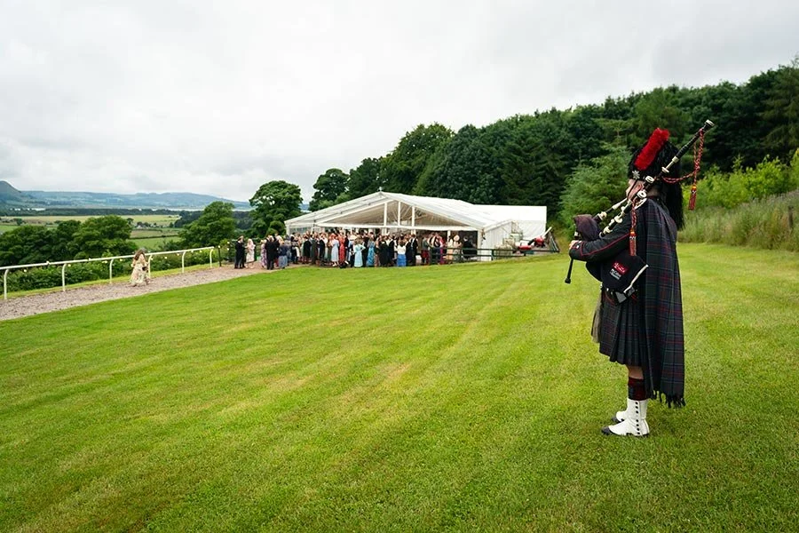 A bagpiper dressed in traditional Scottish attire playing bagpipes on a grassy hill overlooking a wedding reception tent and guests.