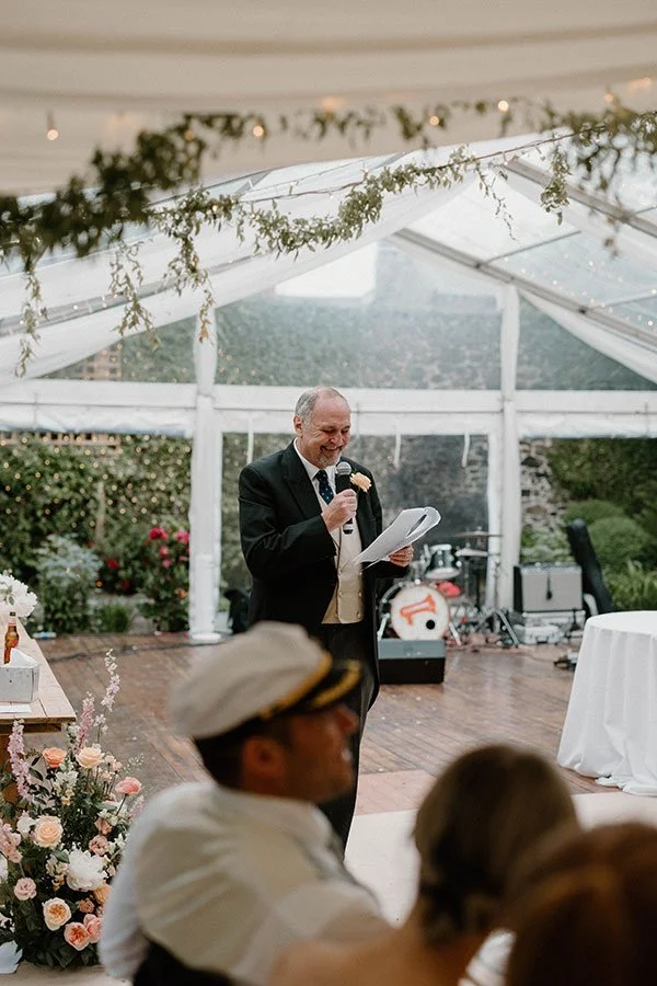 A man in a black suit giving a speech at a wedding reception inside a decorated tent with floral arrangements and string lights.