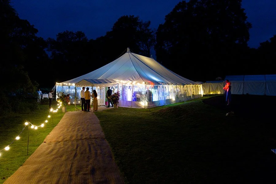 A nighttime outdoor event with a large illuminated white tent, guests inside and outside, and string lights along a pathway leading to the tent.