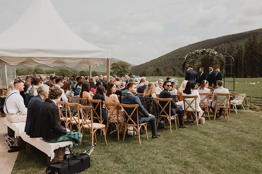 Attending a wedding ceremony outdoors on a grassy field with a large white tent, chairs arranged for guests, a floral arch for the couple, and scenic mountain background.