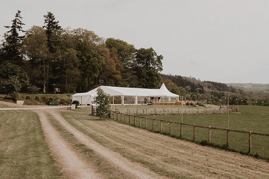Open grassy field with a dirt path leading to a large white event tent, surrounded by wooden fencing and trees, with hills in the background.