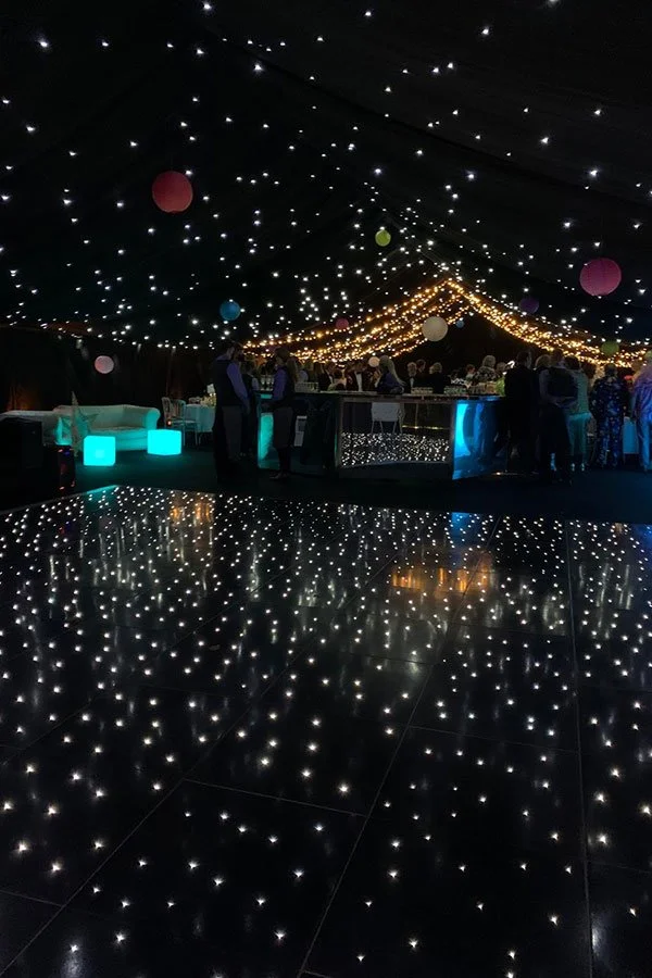 Scene inside a decorated event tent with star-like string lights on the ceiling, hanging paper lanterns, and a crowd of people gathered around a bar or DJ area, with reflective flooring mirroring the ceiling lights.
