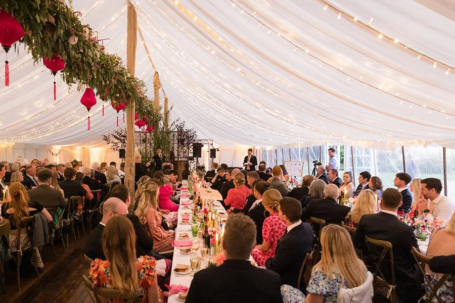 A large outdoor wedding reception under a white tent with string lights and hanging flower arrangements. Guests seated at decorated tables are listening to speakers at a podium.