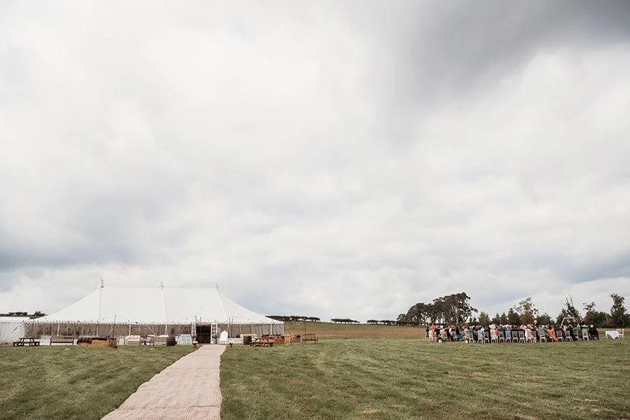 A large white event tent set up outdoors on a grassy field with a pathway leading to it, and a group of people gathering under trees to the right, all under a cloudy sky.