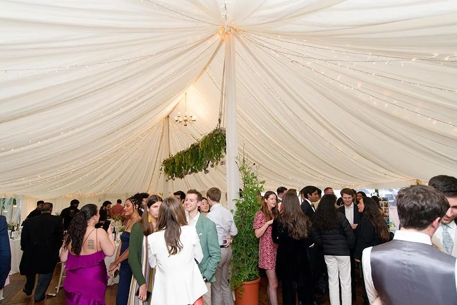 People socializing under a white tent decorated with string lights, with greenery hanging from the ceiling, at a festive event or celebration.