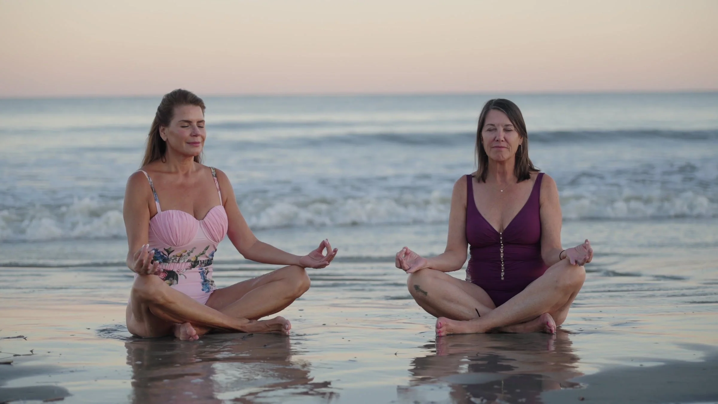 Two women meditating at the water's edge in soft evening light