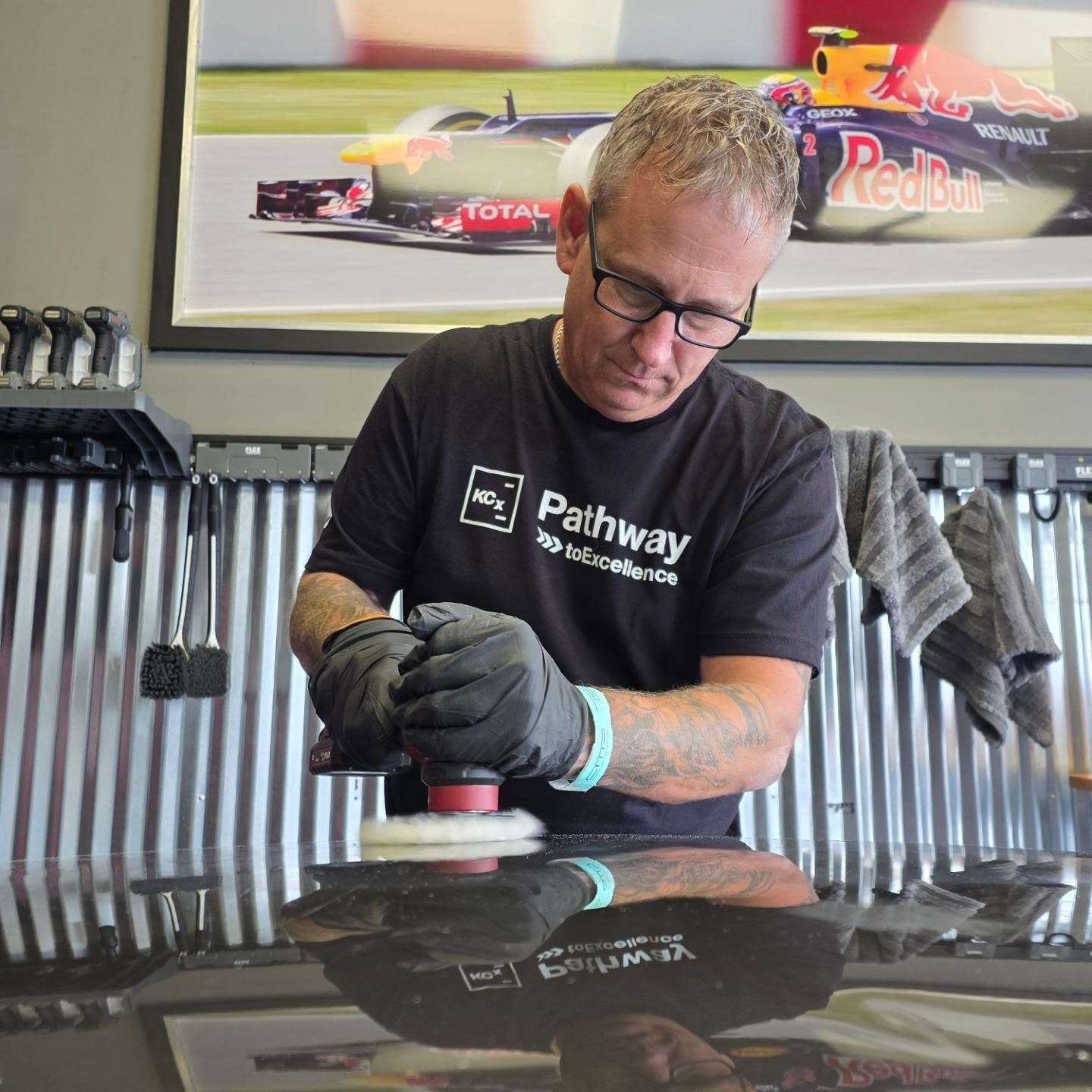Man with glasses and tattoos cleaning a car with a buffer, wearing a black t-shirt with the words "Pathway to Excellence." in front of a large racing poster featuring a Red Bull racing car.