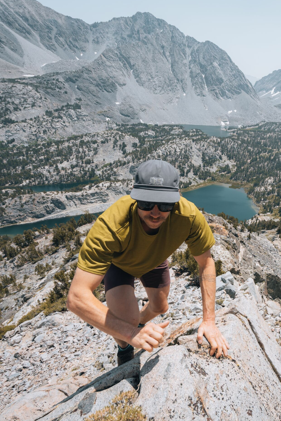 A man wearing a gray cap, Oboz boots, a yellow T-shirt, and Patagonia shorts is climbing on rocky terrain in a mountainous area with lakes and forested slopes in the background.
