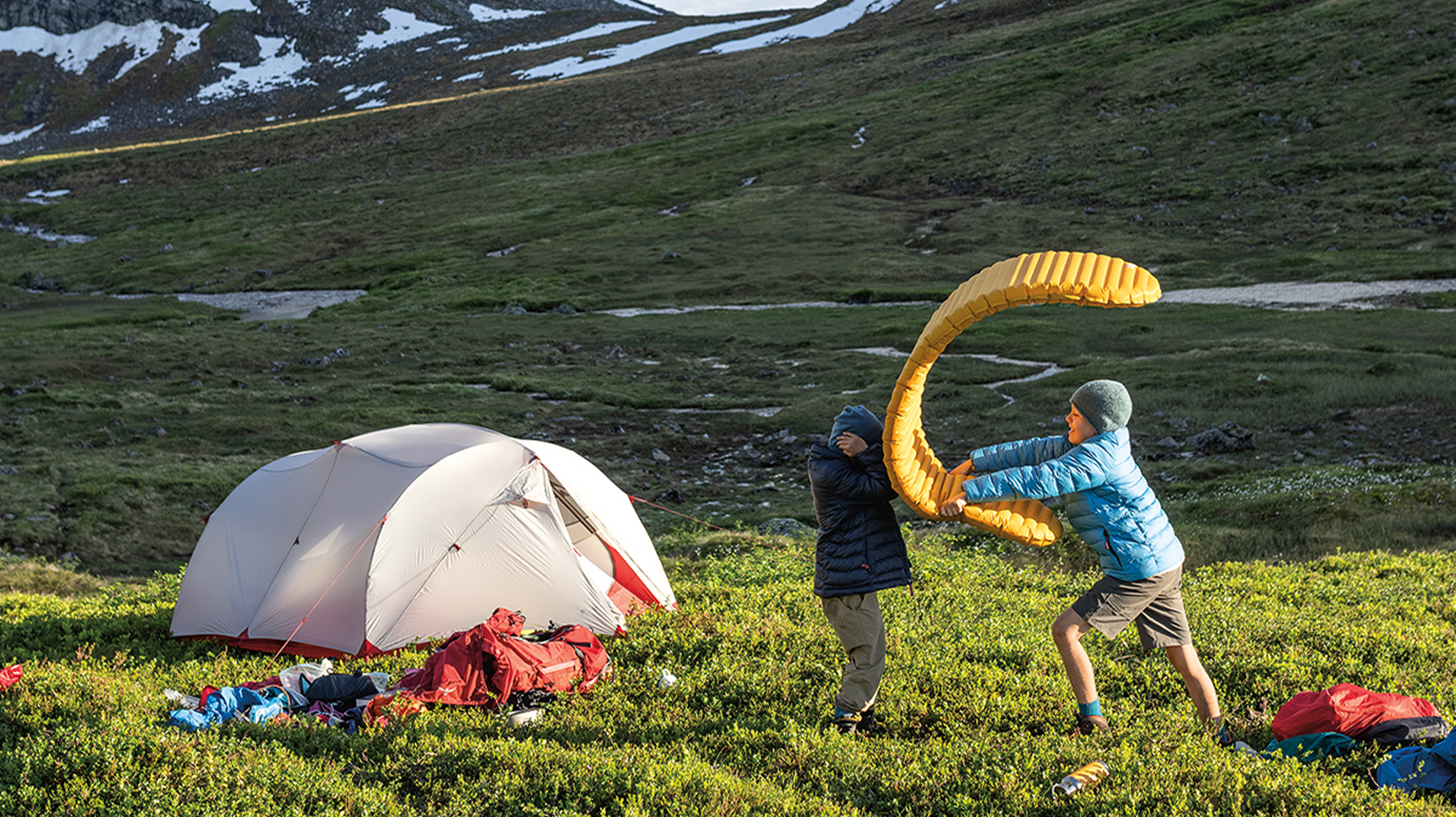Two children playing with a yellow camping foam pad near a Big Agnes tent in a grassy mountain area near Bozeman and Yellowstone National Park.
