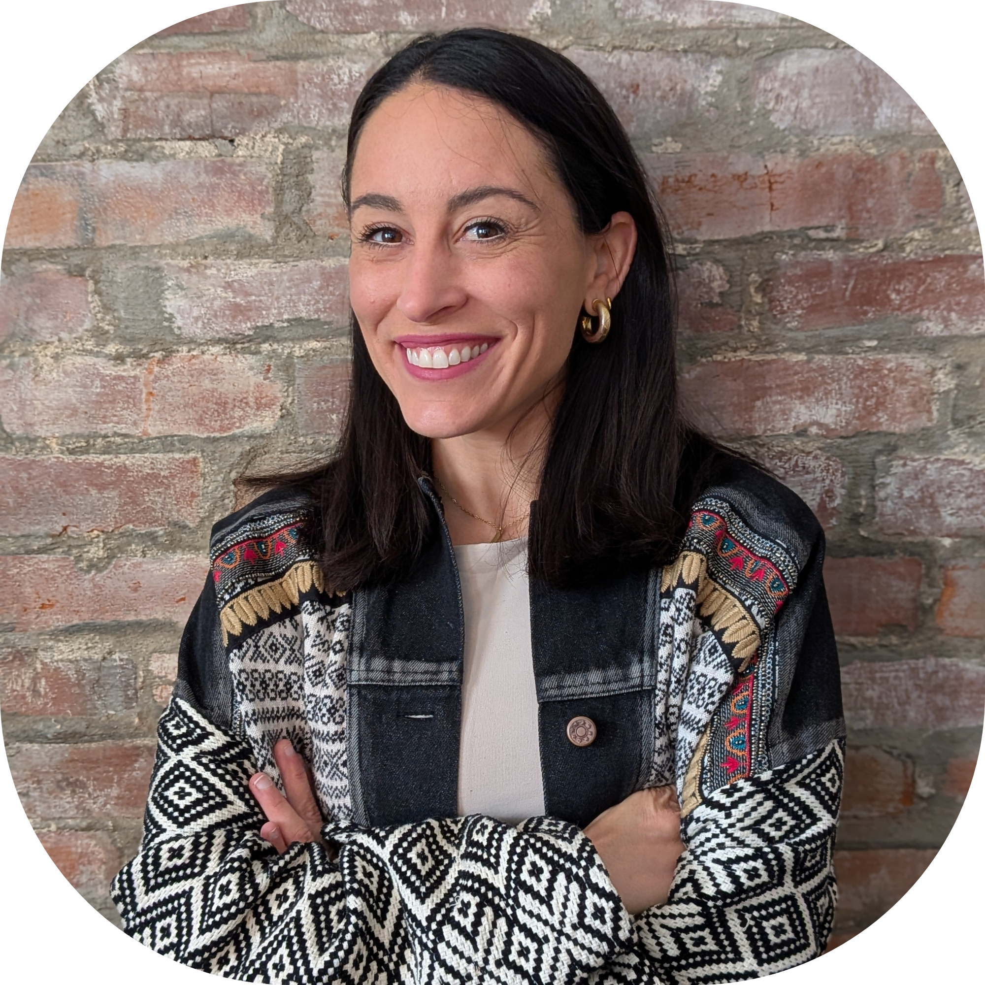 A woman with long dark hair, light skin, smiling, wearing colorful earrings and a multicolored patterned jacket, standing against a brick wall.