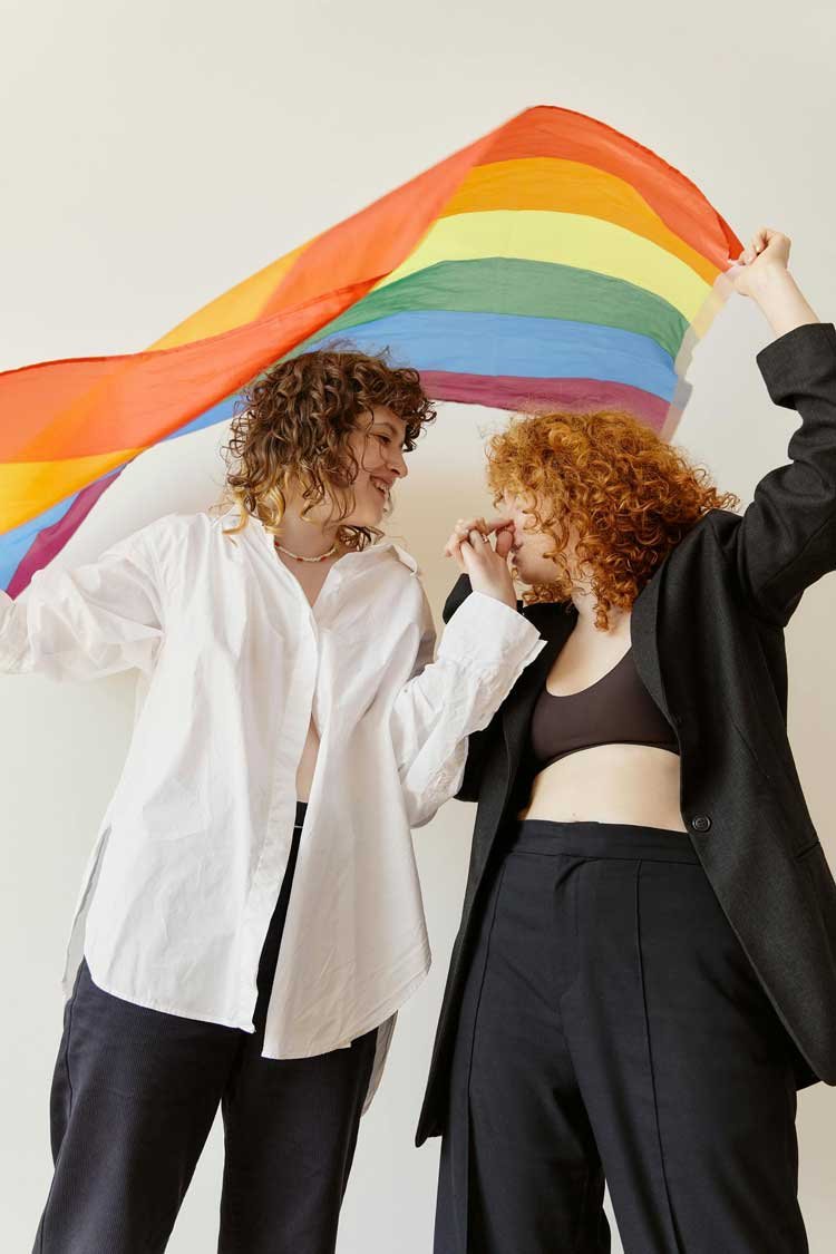Two queer women with curly hair, one with white shirt and the other with black blazer, holding a rainbow flag during a celebration or pride event.