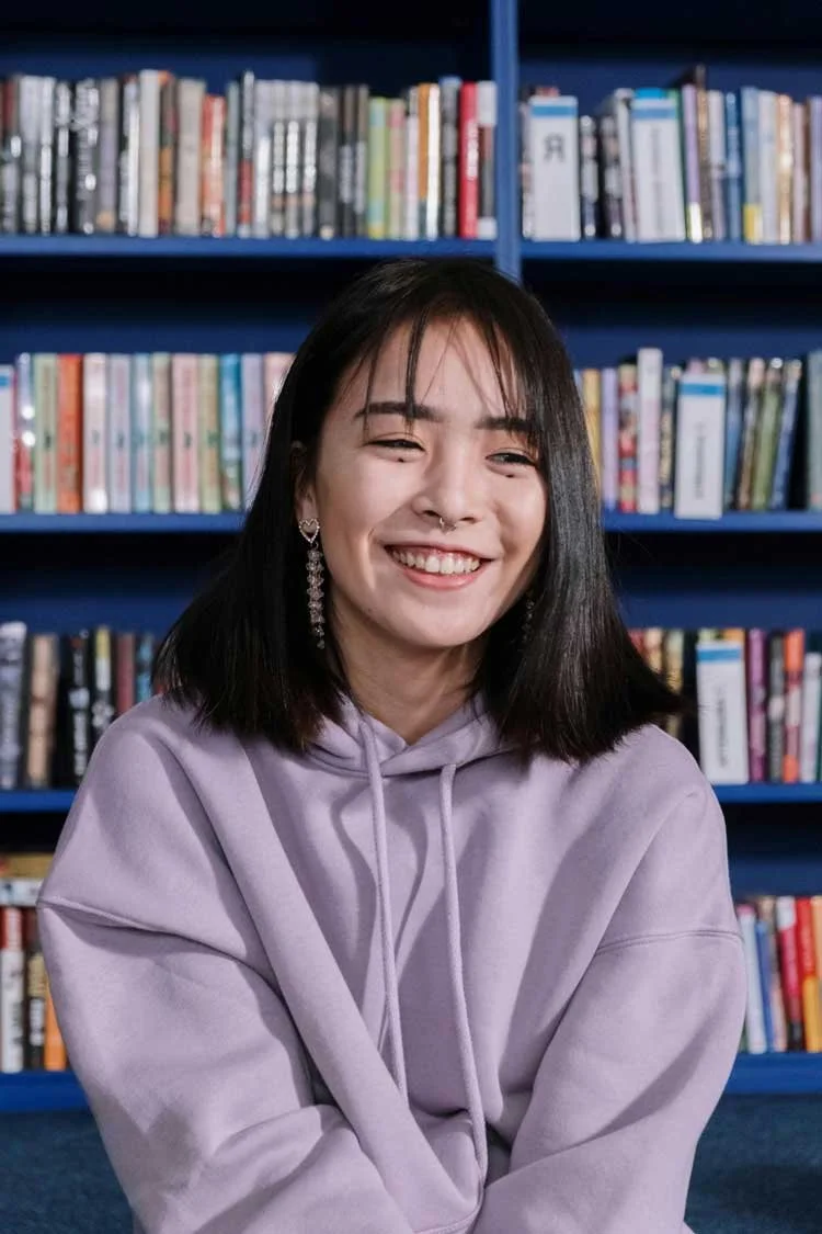 A young Asian woman with shoulder-length black hair, wearing a lavender hoodie and earrings, smiling at the camera in front of a bookshelf filled with books.