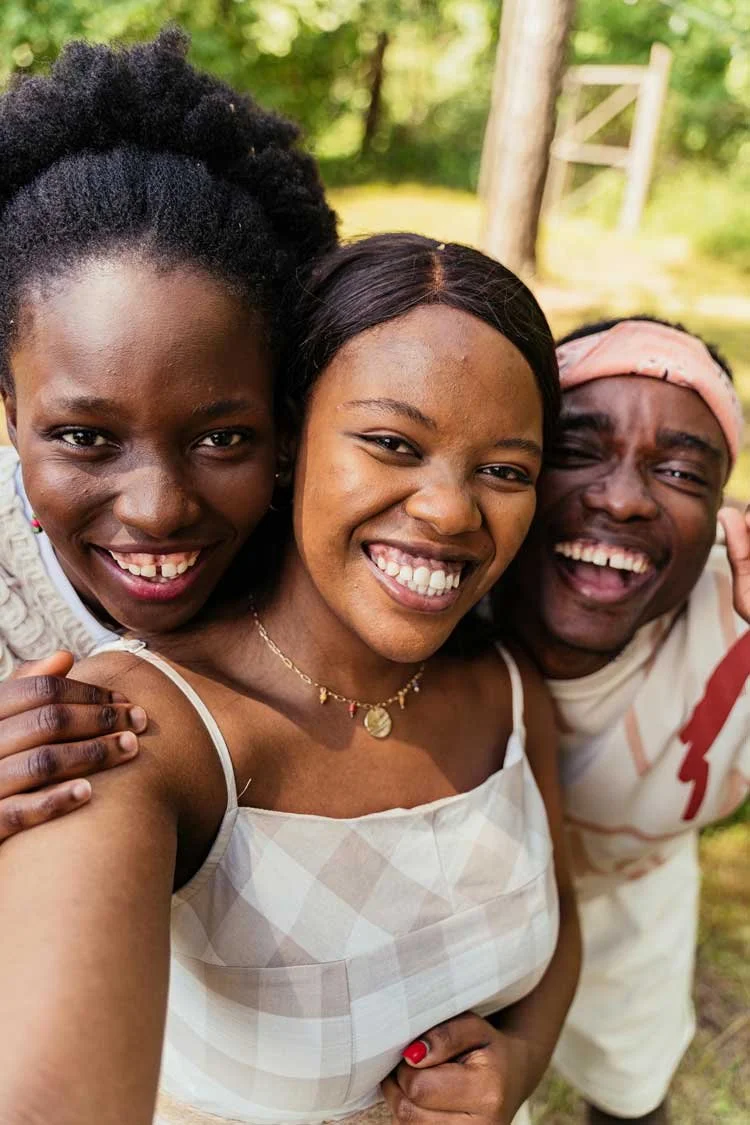 Three smiling millenial adults taking a selfie outdoors in a green, wooded area.
