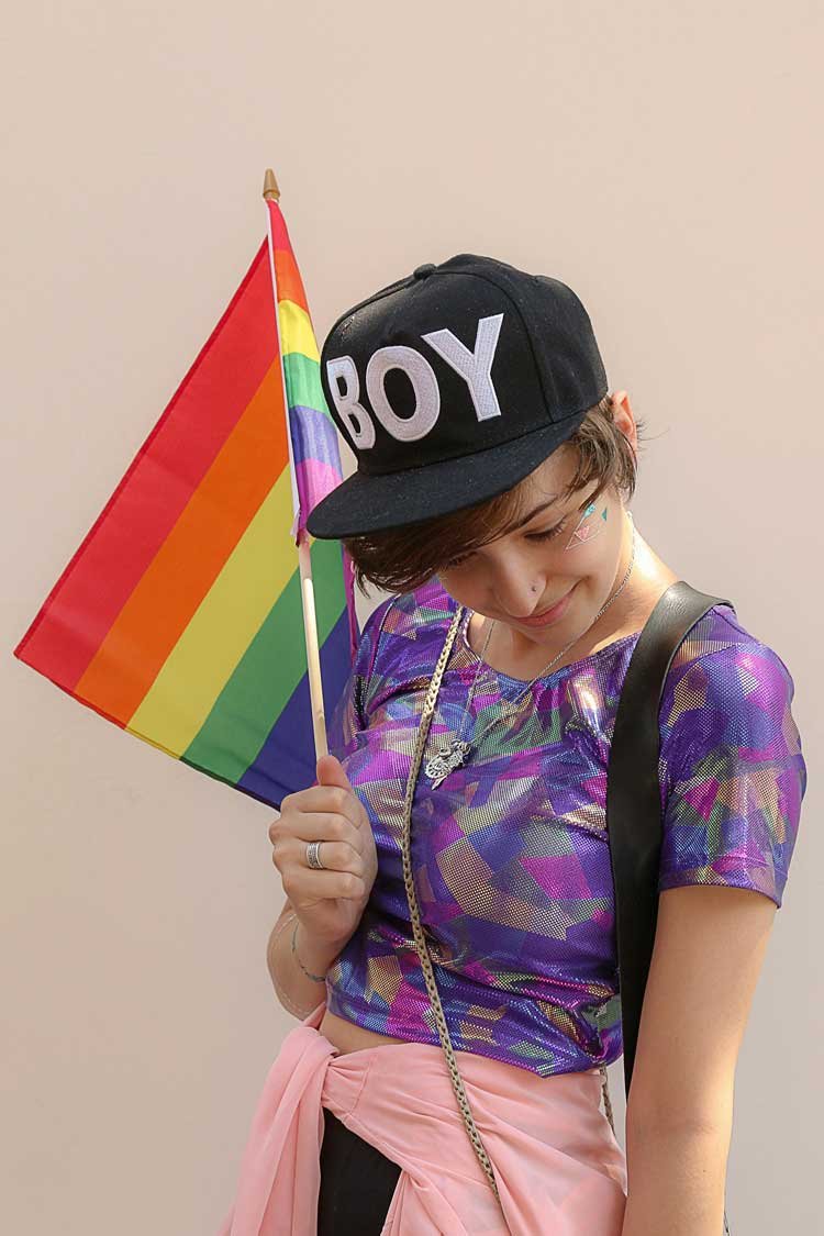 A person holding a rainbow pride flag, wearing a black cap with the word 'BOY', a colorful shiny top, pink jacket tied around the waist, and colorful face stickers.
