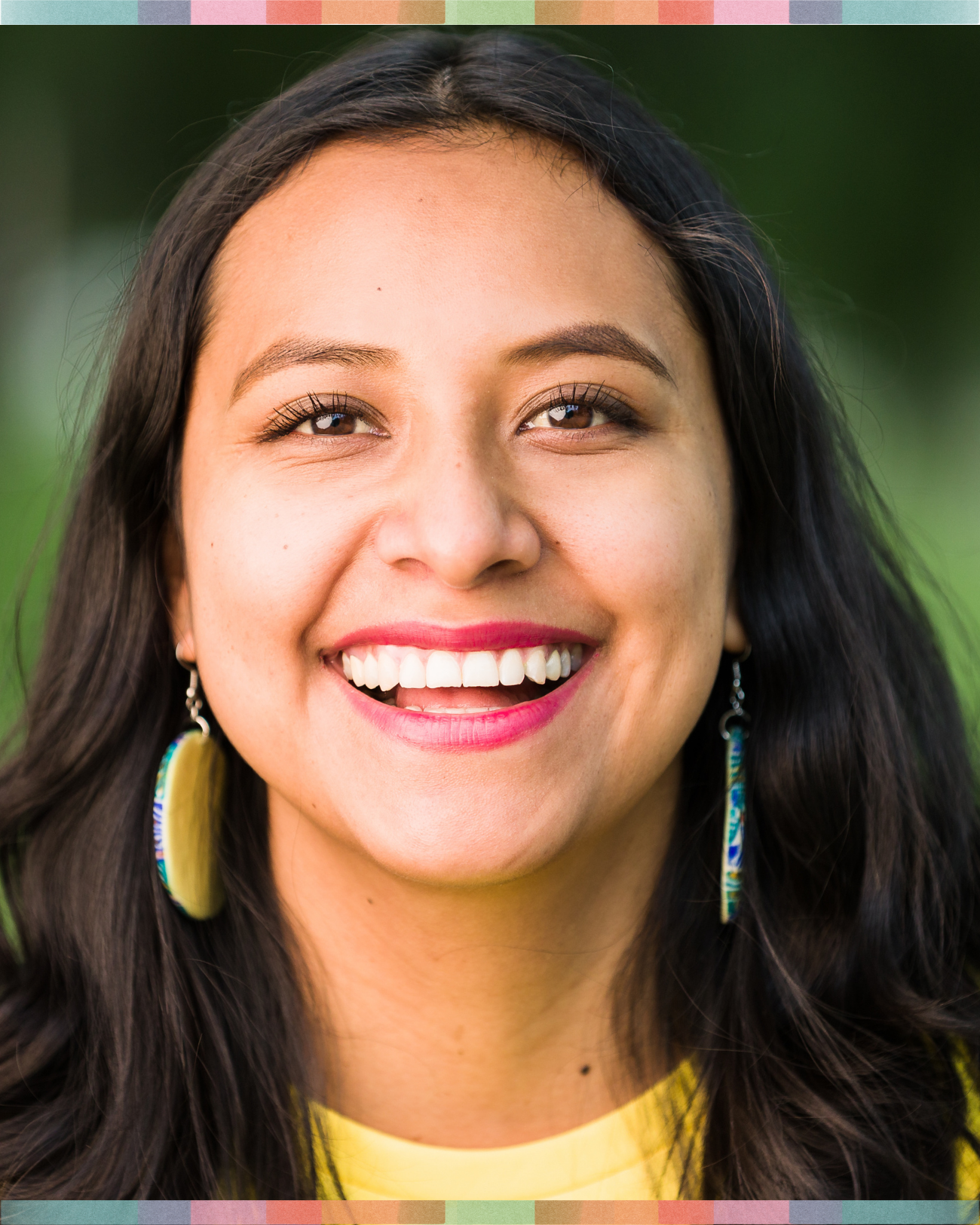 Close-up of a smiling woman with long dark hair, wearing colorful earrings and a yellow top, outdoors with a green blurred background.