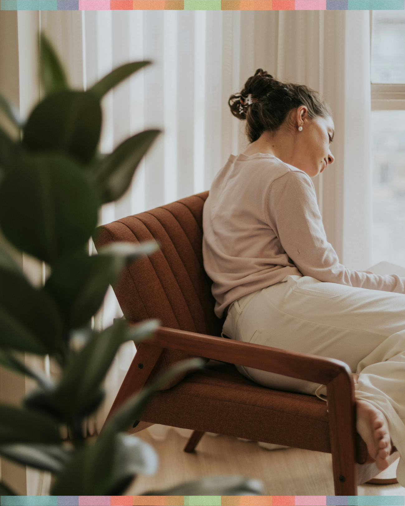 A woman sitting on a wooden chair with a reddish-brown cushion, leaning back with her eyes closed, near a window with sheer white curtains. A large green plant is in the foreground.