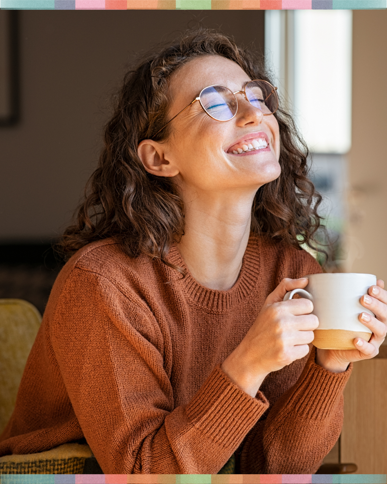A woman with curly brown hair and glasses smiling while holding a beige and white mug.