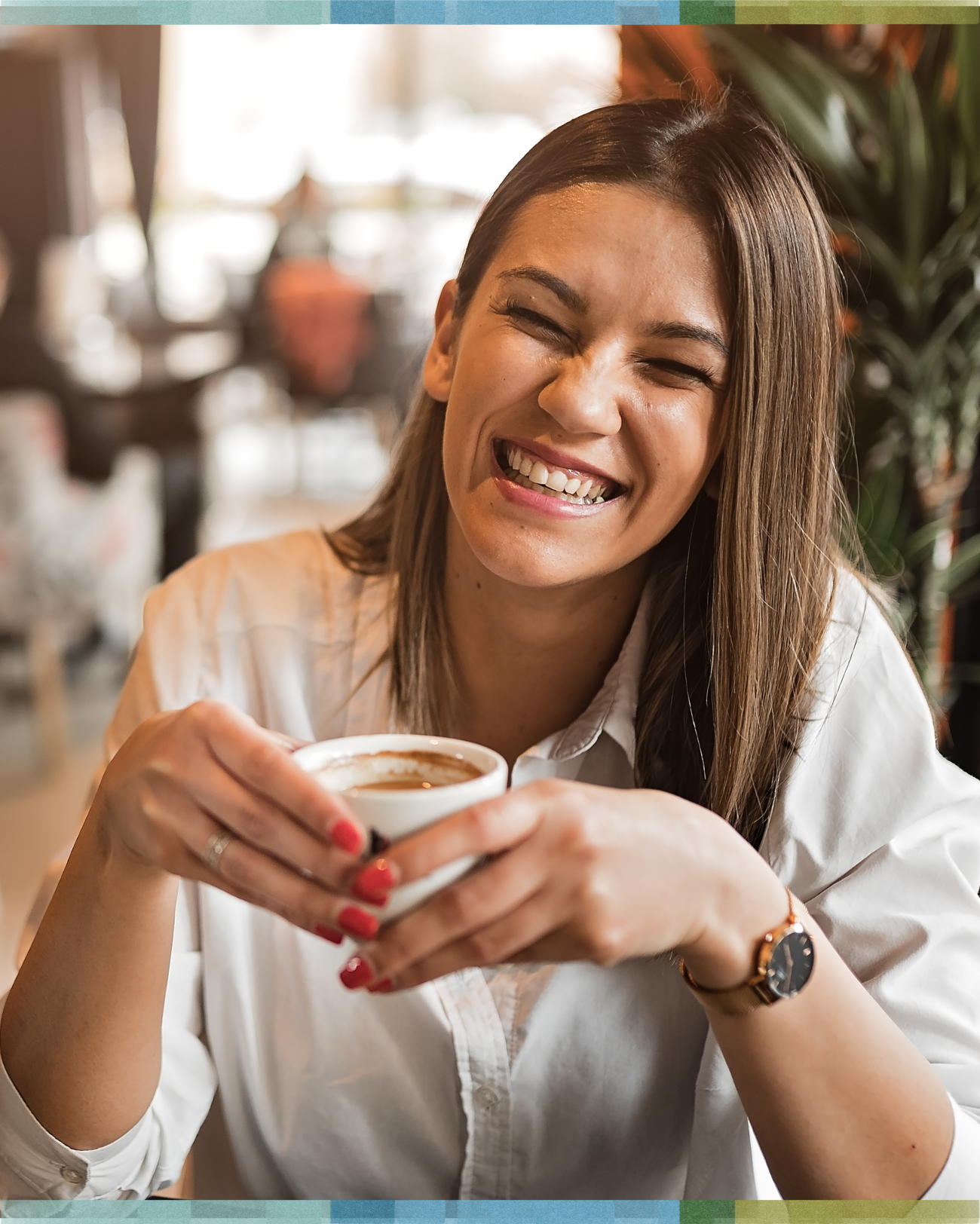 A woman smiling with eyes closed, holding a cup of coffee inside a cozy cafe, with blurred background and greenery.